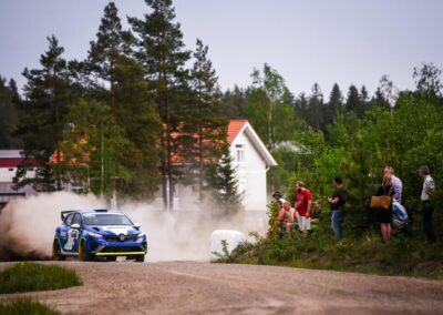 A Renault rally car speeds past spectators, leaving a cloud of dust in its wake.